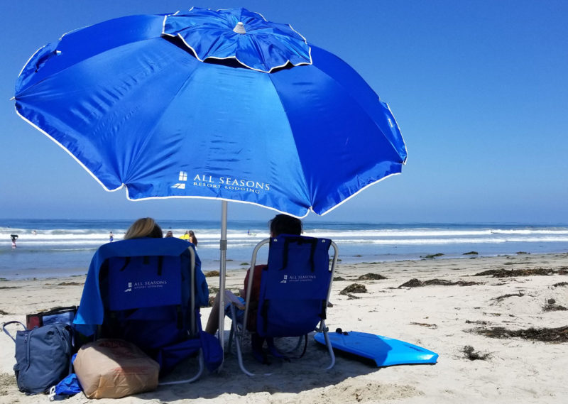 Two Beach Bathers Resting Under a Blue All Seasons Resort Lodging Umbrella on a Sunny Day